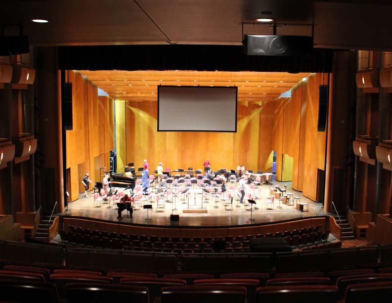 Orchestra stage setup in a concert hall with chairs, music stands, and a projection screen above.